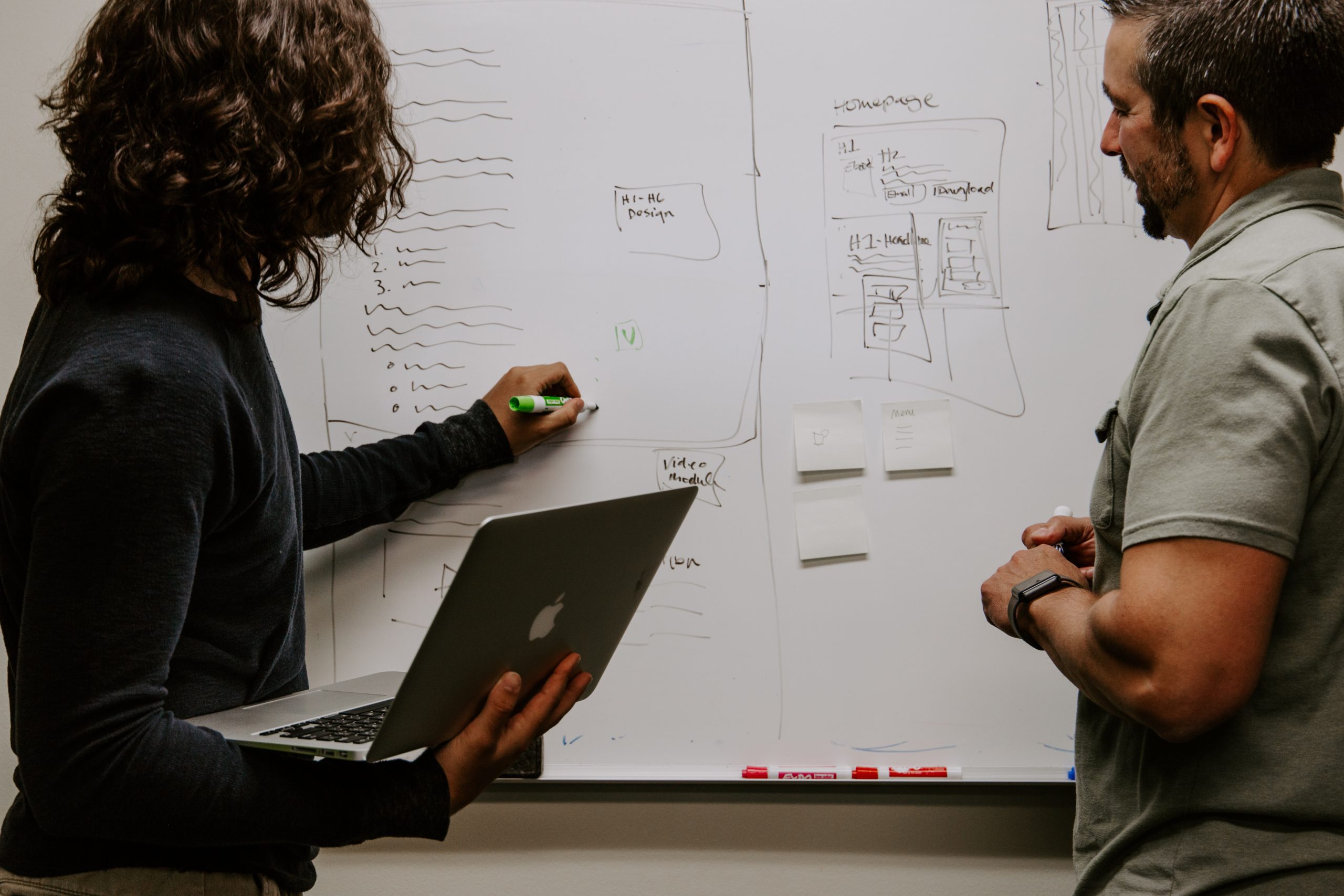 A woman writes on a whiteboard while holding a computer as a man looks on.